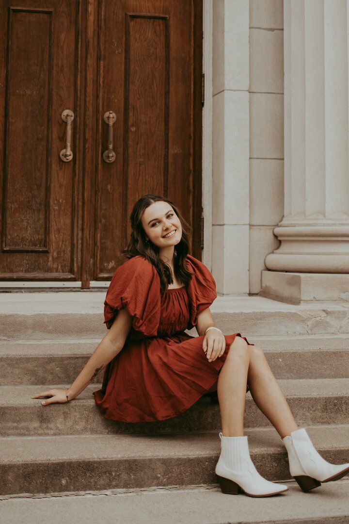 Smiling woman in a red dress and white boots sitting on stone steps near wooden doors.